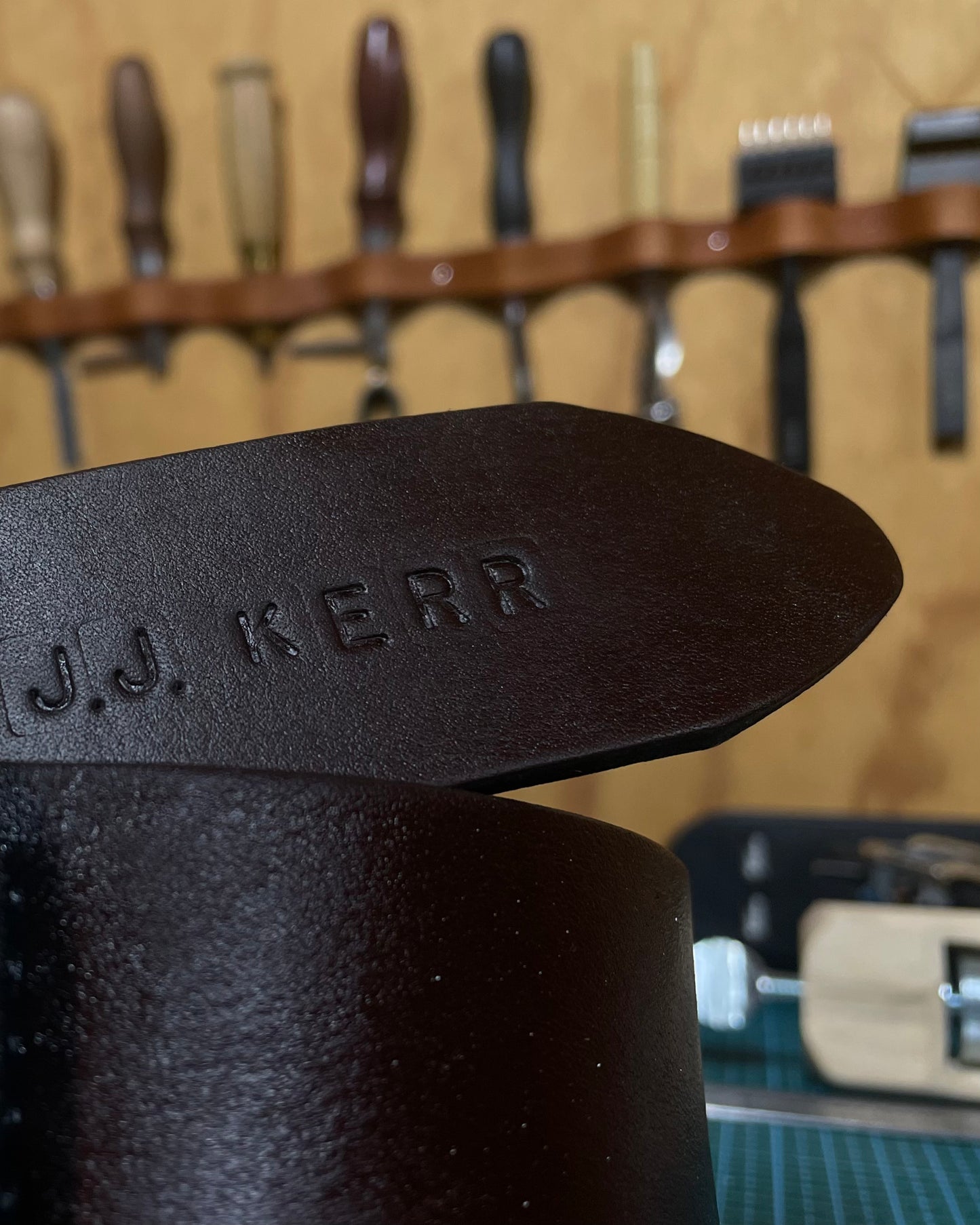 Close-up of a brown leather belt with 'J.J. Kerr' custom embossing, set against a blurred background of tools.