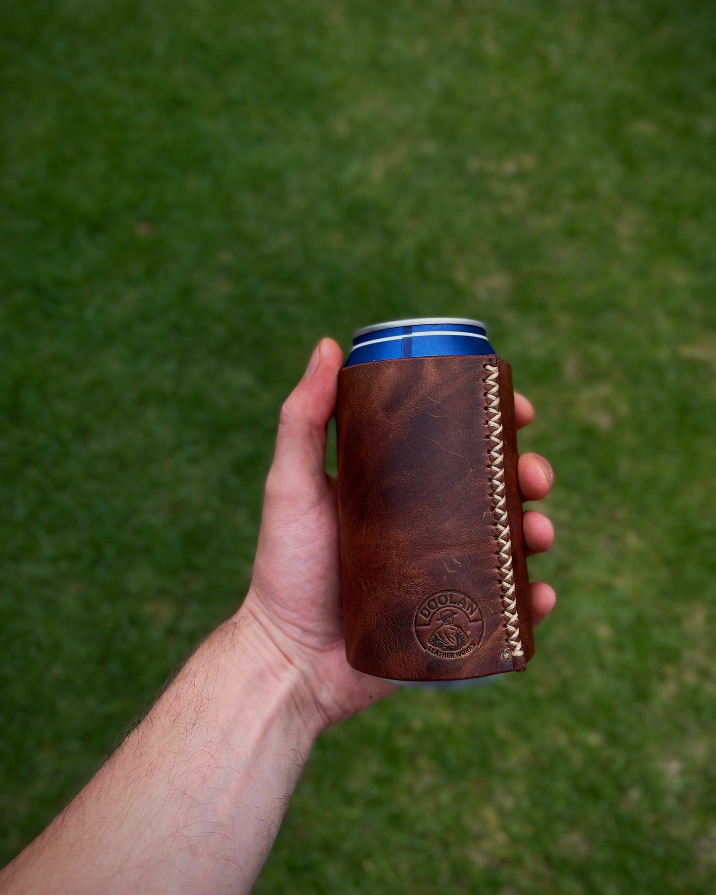 Hand holding a brown leather can cooler with a green grass background