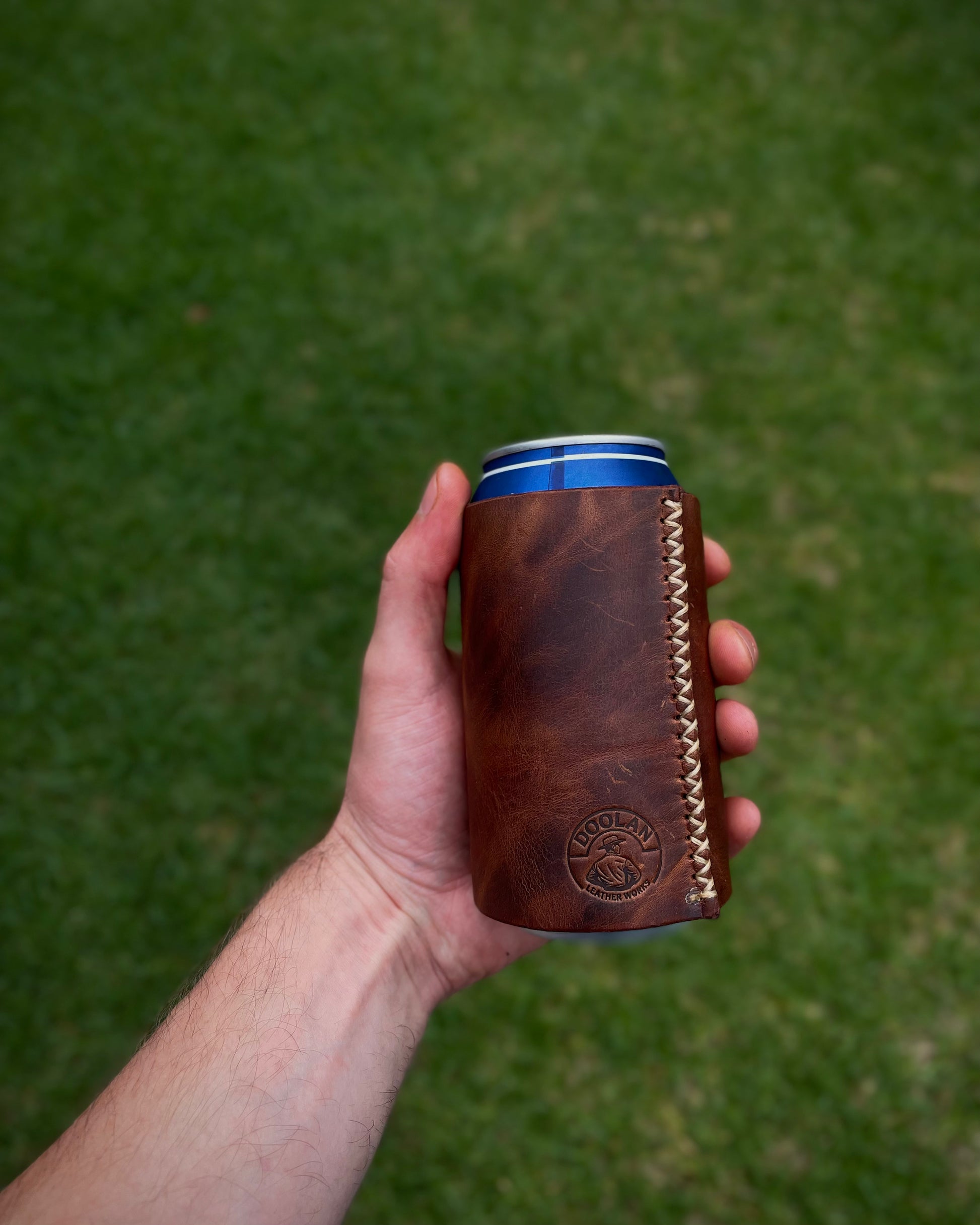 Hand holding a brown leather can cooler with a green grass background
