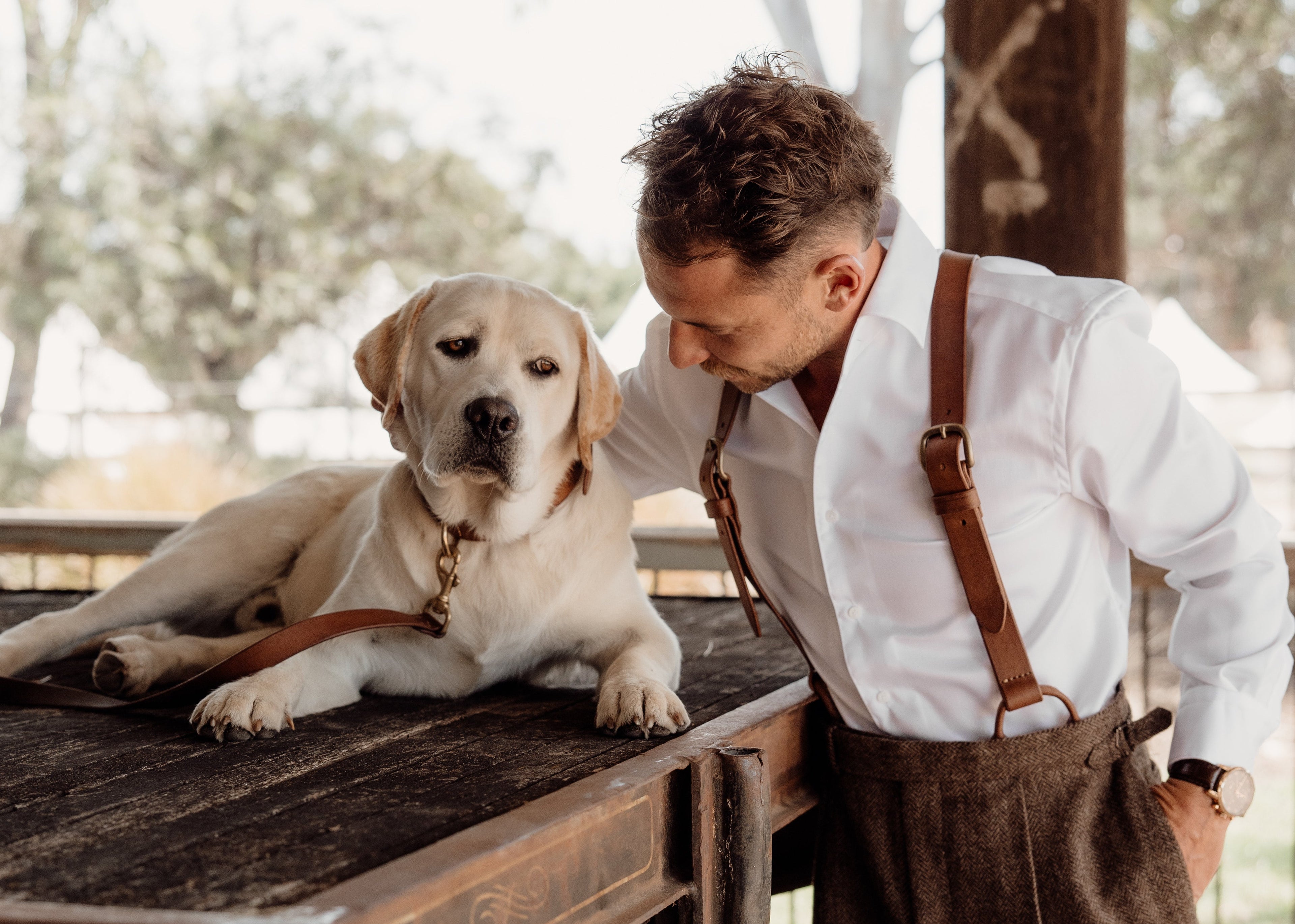 Man in suspenders standing next to a dog on a wooden platform outdoors