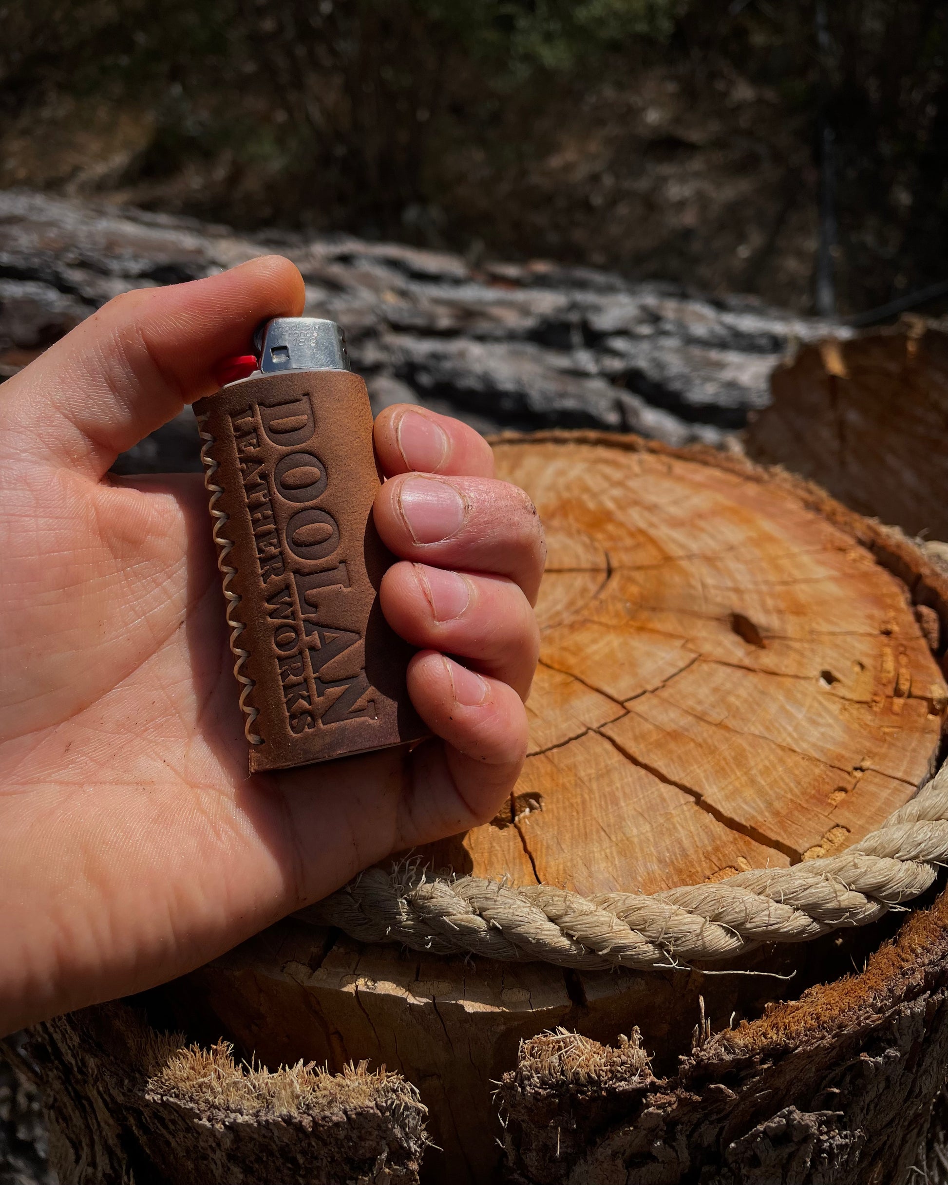 Hand holding a leather lighter with 'Doolan Leather Works' branding on a wooden log background.