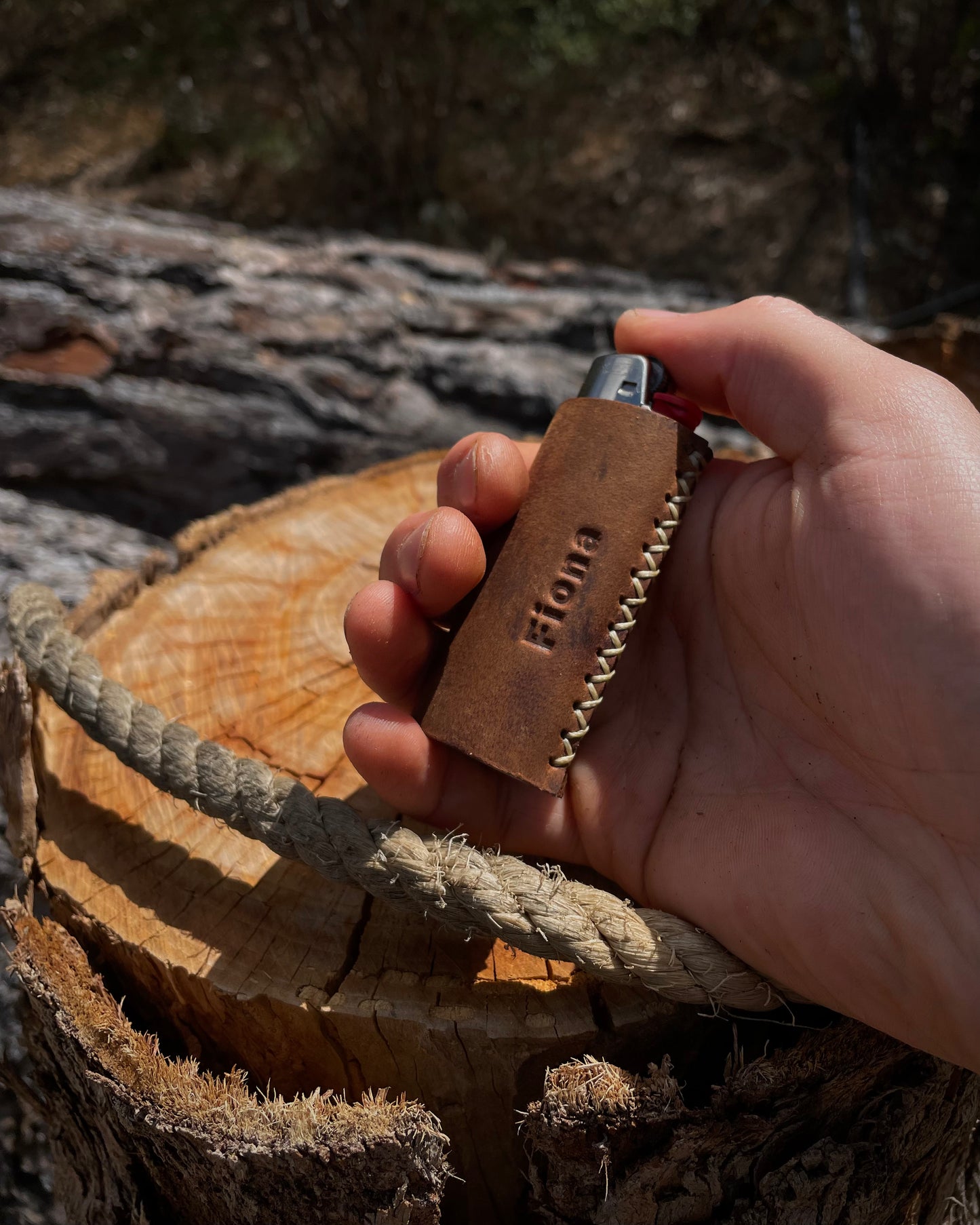 Hand holding a leather lighter case with 'Flona' branding on a wooden log background
