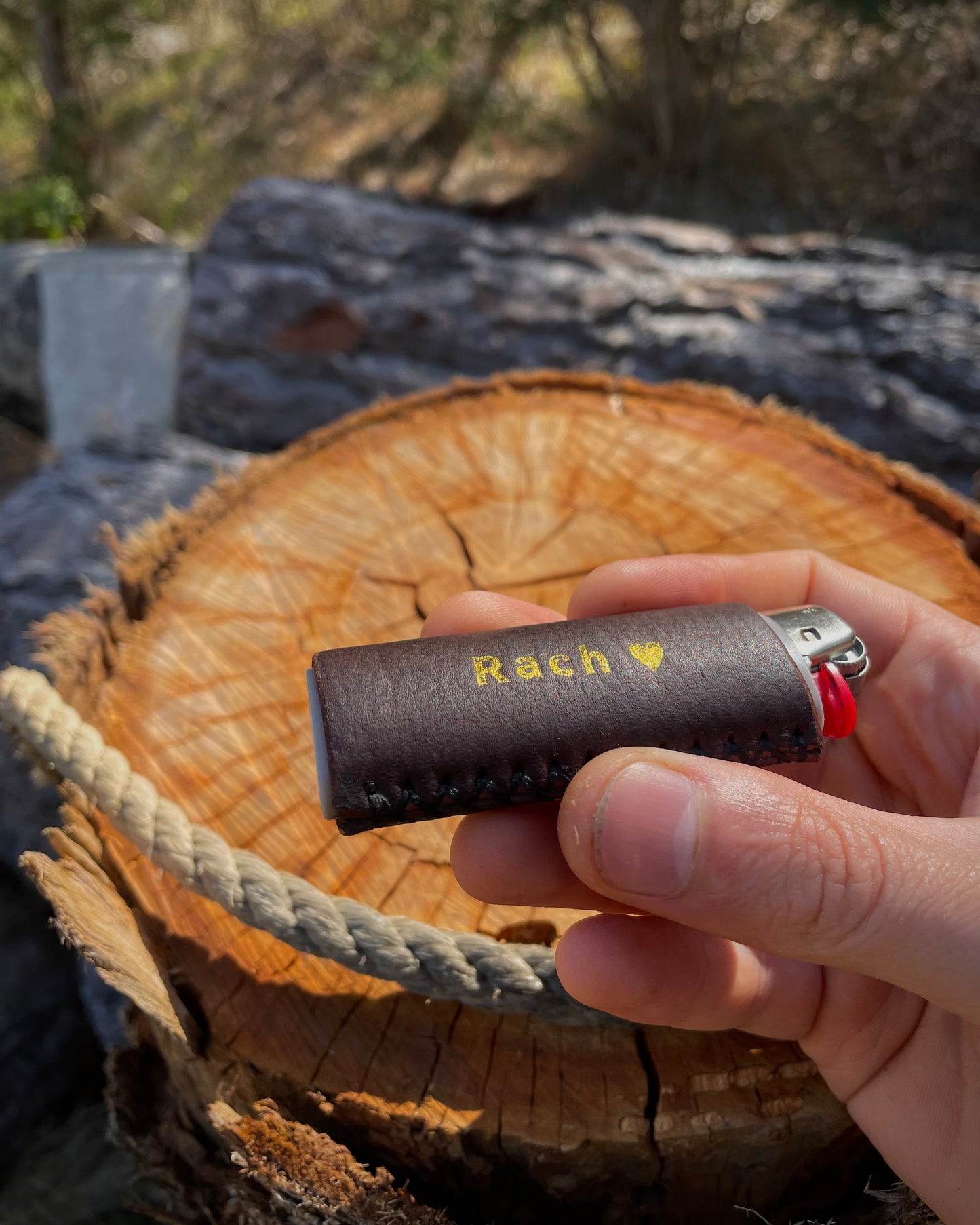 Hand holding a brown leather lighter case with 'Rach' engraved on it, against a natural background.