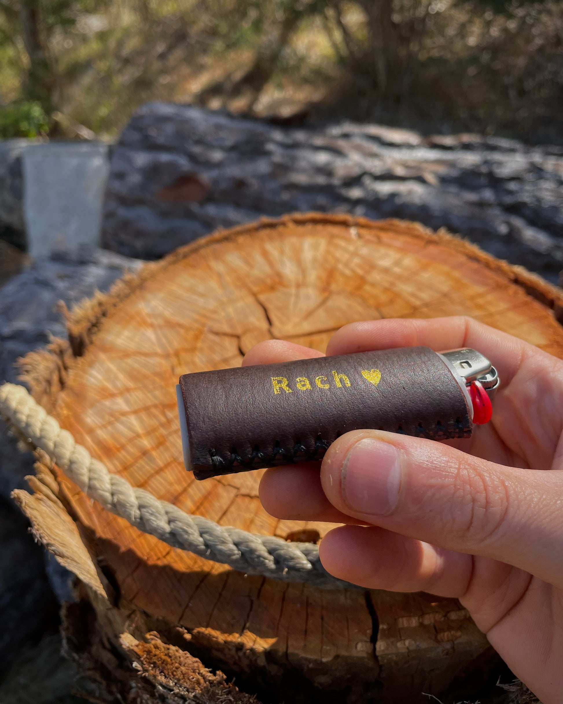 Hand holding a brown leather lighter case with 'Rach' engraved on it, against a natural background.
