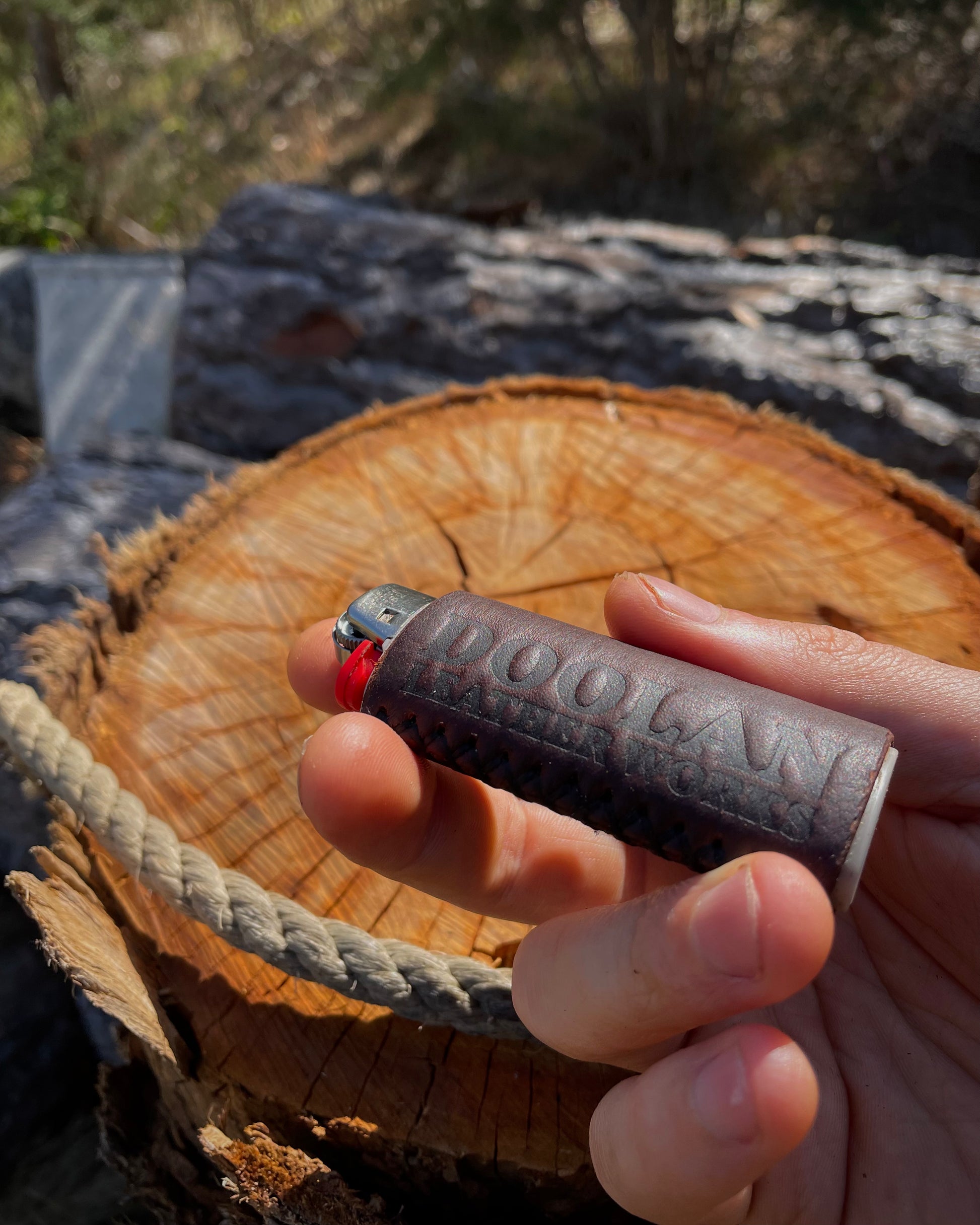 Hand holding a brown leather lighter cover on a wooden log with a natural outdoor background