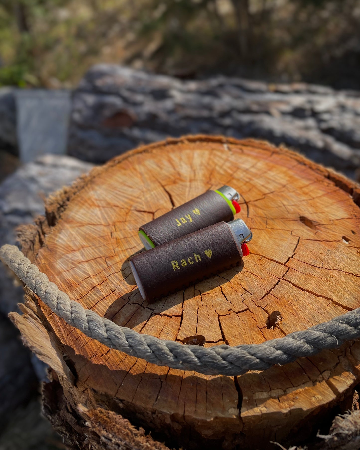 Two leather lighter cases on a wooden log with 'Jay' and 'Rach' engraved, surrounded by natural elements.