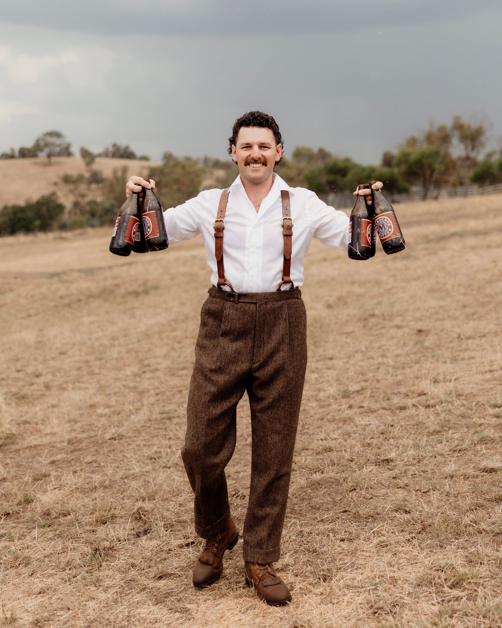 Man in suspenders holding beer bottles in a field