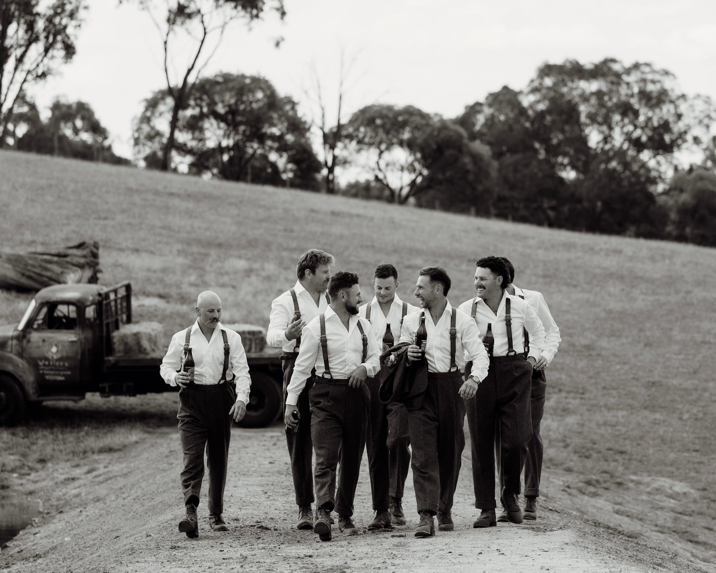 Group of men in suspenders standing on a dirt road with a truck in the background.