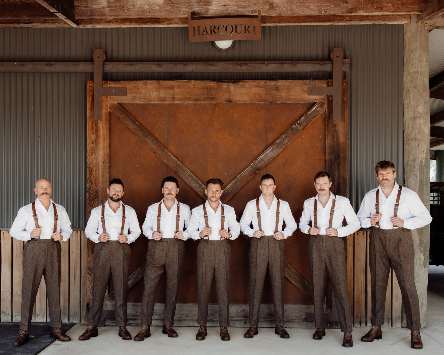 Seven men in formal attire with suspenders standing in front of a rustic wooden door.