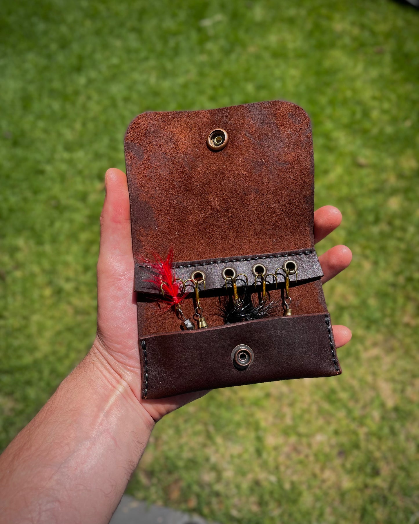 Brown leather fishing lure case held in a hand with grass background
